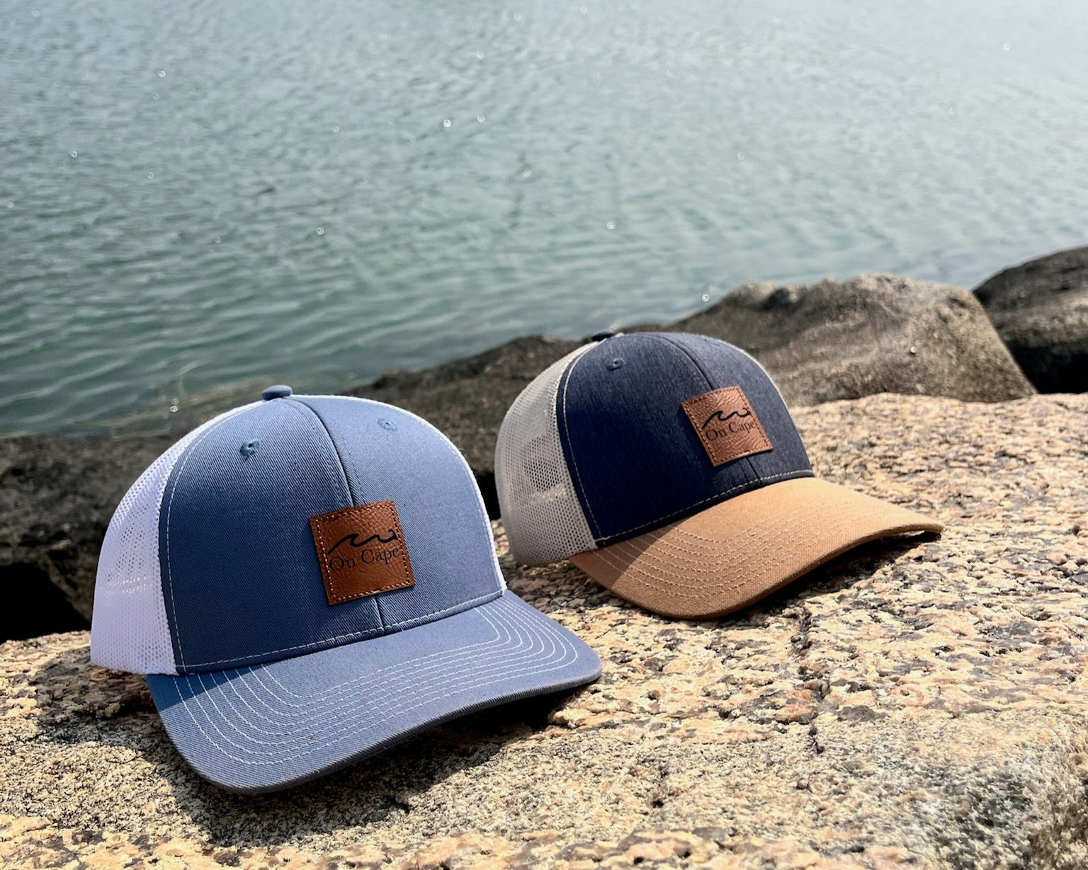 On Cape Trucker Hats with leather patches on a stone surface by a body of water.