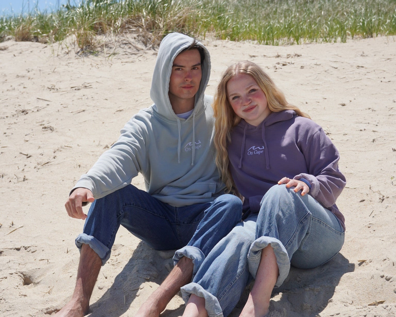Two people sitting on a sandy beach wearing matching hoodies and jeans.